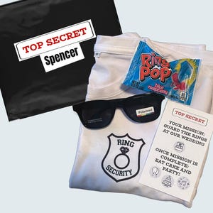 May include: A white t-shirt with a black ring security badge, sunglasses, a package of Ring Pop candy, and a note that says "Top Secret: Your Mission: Guard the Rings at Our Wedding. Once Mission is Complete: Eat Cake and Party!"