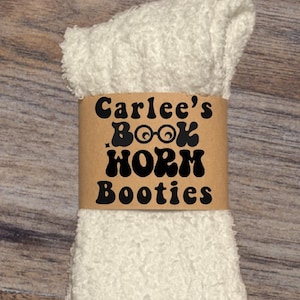 May include: A pair of white, fluffy booties with a brown paper band. The band features the text "Carlee's Book Worm Booties" in a black, playful font. The booties appear soft and cozy.