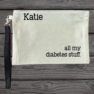 May include: Cream-colored canvas pouch with a black zipper and wrist strap. The text "Katie" is printed at the top, with "all my diabetes stuff" below. The pouch is rectangular and sits on a wooden surface.