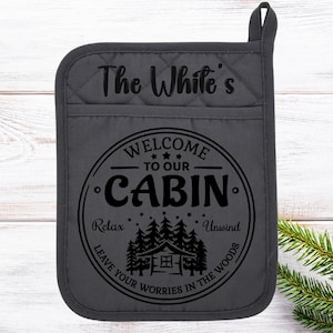 May include: A dark gray quilted pot holder with a pocket. The text "The White's" is at the top. Below is a circular design that reads "Welcome to our Cabin" with a cabin and trees graphic. The words "Relax" and "Unwind" are also included.