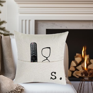 May include: A white pillow with a pocket on the front. The pocket contains a remote control and a pair of glasses. The pillow has the letter "S." printed on the front.