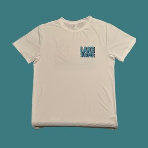 May include: White t-shirt with a blue graphic that says "LAKE SURFER" on the front.