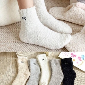 May include: White fuzzy socks with a small black bow embroidered near the cuff. The image also shows several pairs of similar socks in various colors, including cream, gray, and black. The socks appear soft and comfortable, suitable for lounging.