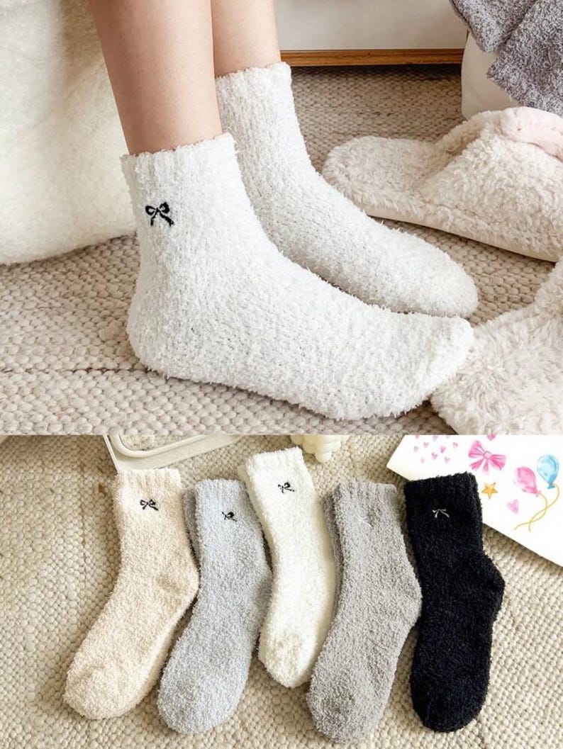 May include: White fuzzy socks with a small black bow embroidered near the cuff. The image also shows several pairs of similar socks in various colors, including cream, gray, and black. The socks appear soft and comfortable, suitable for lounging.