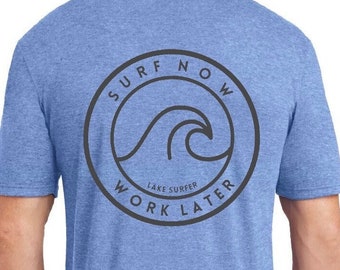May include: A blue heather t-shirt with a black circle graphic on the back. The graphic says "Surf Now Work Later" with a wave design inside the circle. The text "Lake Surfer" is written in smaller font below the wave.