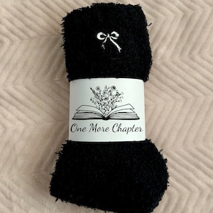 May include: Black fuzzy socks with a white bow detail and a white band. The band features an open book graphic with floral accents and the text "One More Chapter". The socks are rolled up and ready for gifting.