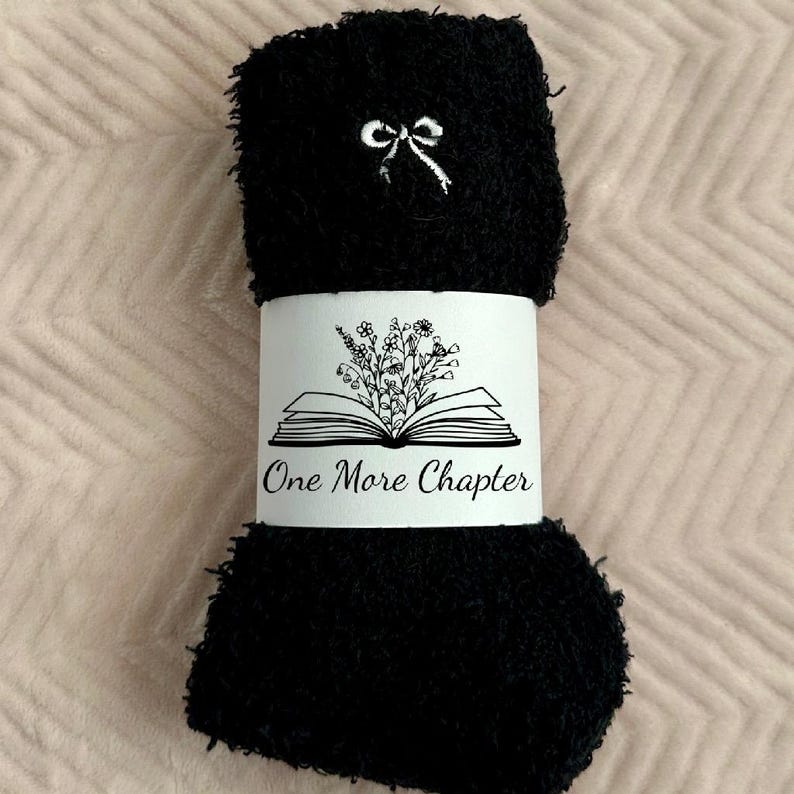 May include: Black fuzzy socks with a white bow detail and a white band. The band features an open book graphic with floral accents and the text "One More Chapter". The socks are rolled up and ready for gifting.