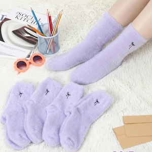 May include: A pair of lavender fuzzy socks with a small black bow embroidered on the side, along with three additional pairs. The socks are displayed on a white, textured surface. A pair of sunglasses and pencils are also visible.