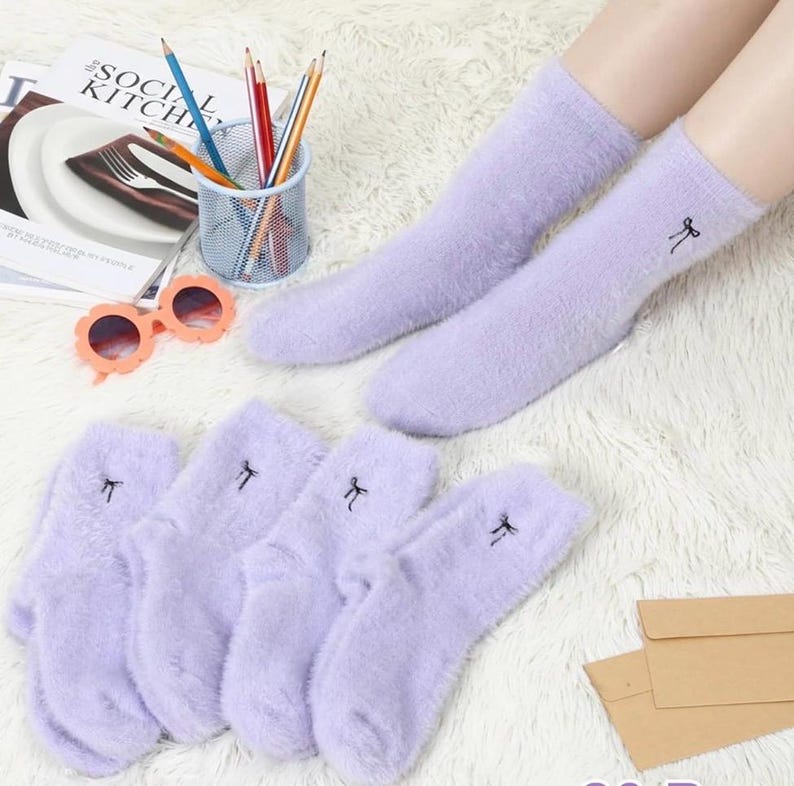 May include: A pair of lavender fuzzy socks with a small black bow embroidered on the side, along with three additional pairs. The socks are displayed on a white, textured surface. A pair of sunglasses and pencils are also visible.