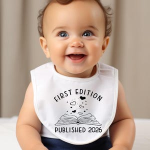 May include: A white baby bib with the text "FIRST EDITION PUBLISHED 2026" in black. The bib features an open book design with hearts and stars. The baby is smiling.