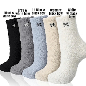 May include: A collection of knee-high fuzzy socks in various colors: black with a white bow, gray with a white bow, light blue with a black bow, cream with a black bow, and white with a black bow. Each sock has a bow detail.
