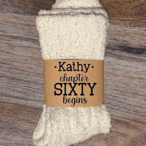 May include: A pair of white, fluffy socks wrapped with a brown paper band. The band features the text "Kathy chapter SIXTY begins" in a black, bold font. The socks are folded and appear soft.