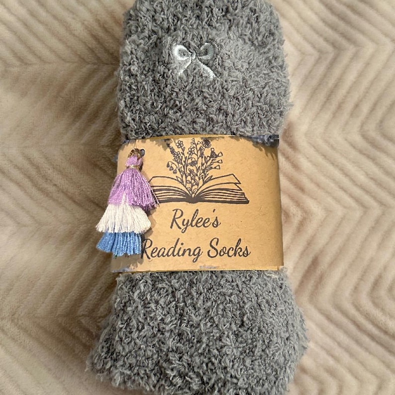 May include: Gray fuzzy socks with a small white bow and a decorative tassel. The socks are wrapped with a brown paper band that reads "Rylee's Reading Socks" with a floral and book design.