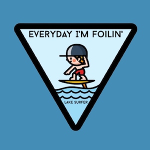 May include: A black and white illustration of a person wearing a blue hat and red shorts, riding a surfboard on a blue wave. The text "EVERYDAY I'M FOILIN'" is above the person and the text "LAKE SURFER" is below the person.