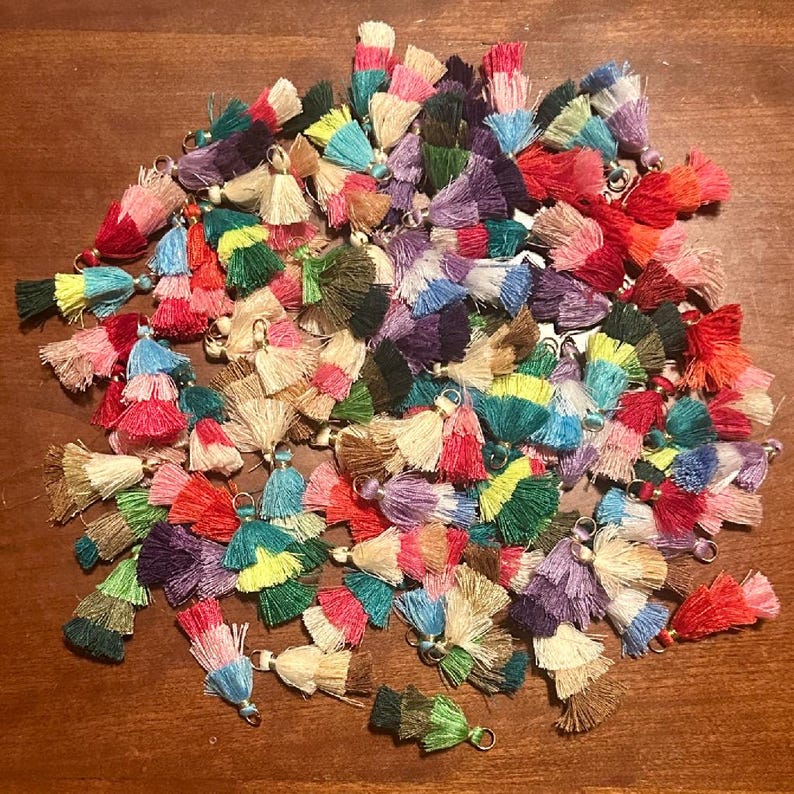 May include: A collection of colorful tassels in various shades, including red, pink, green, blue, and yellow. Each tassel is made of layered threads and has a small metal loop at the top. The tassels are scattered on a wooden surface.