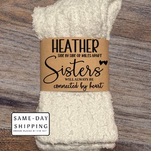 May include: A pair of white fuzzy socks with a brown paper band. The band reads "HEATHER SIDE BY SIDE OR MILES APART Sisters WILL ALWAYS BE connected by heart."