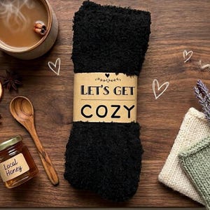 May include: A pair of black fuzzy socks wrapped with a brown paper band that reads "Let's Get Cozy." The image includes a wooden spoon, a jar of honey, a cup of tea, and a knitted item.