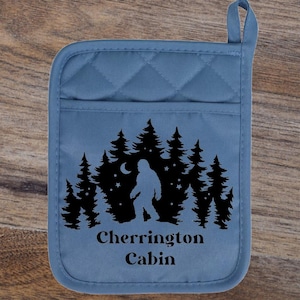 May include: A blue quilted pot holder with a black graphic of a forest, a crescent moon, and a Bigfoot silhouette. The text "Cherrington Cabin" is printed below the graphic. Includes a hanging loop.