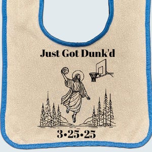 May include: A white bib with a blue trim, featuring a black and white illustration of a person dunking a basketball. The text "Just Got Dunk'd" and "3.25.25" are printed on the bib.