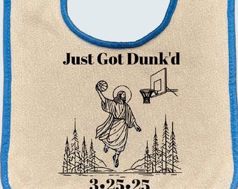 May include: A white bib with a blue trim, featuring a black and white illustration of a person dunking a basketball. The text "Just Got Dunk'd" and "3.25.25" are printed on the bib.