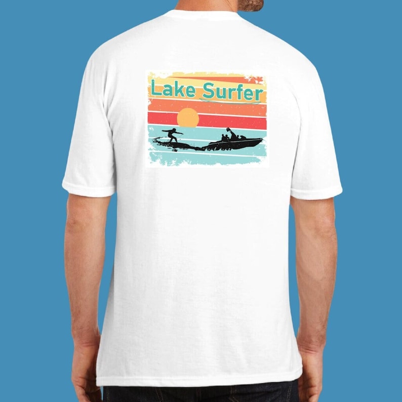 May include: White t-shirt with a vintage-style graphic on the back. The graphic features a sunset with a boat and a person waterskiing. The text "Lake Surfer" is printed in teal above the graphic.