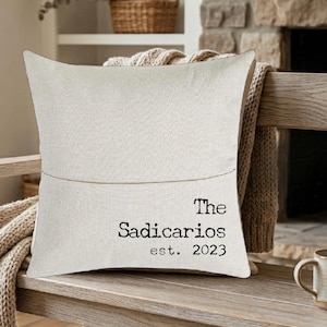 May include: A square off-white linen throw pillow cover featuring a thin horizontal line across the center and black printed text that reads The Sadicarios est. 2023 in a typewriter-style font, centered on the lower right portion of the fabric.