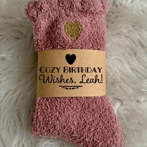 May include: A pair of dusty rose fuzzy socks featuring a small gold embroidered heart. A tan paper band wraps around the center, printed with a black heart icon and the text: COZY BIRTHDAY Wishes, Leah! with a decorative line underneath.