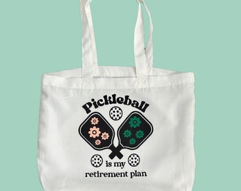 May include: White canvas tote bag with a black and white graphic of two pickleball paddles and the text "Pickleball is my retirement plan".