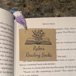 May include: A bookmark with a tassel and a decorative tag. The tag is brown with an illustration of an open book and flowers. The text "Rylee's Reading Socks" is printed on the tag. The tassel is blue, white, and purple.
