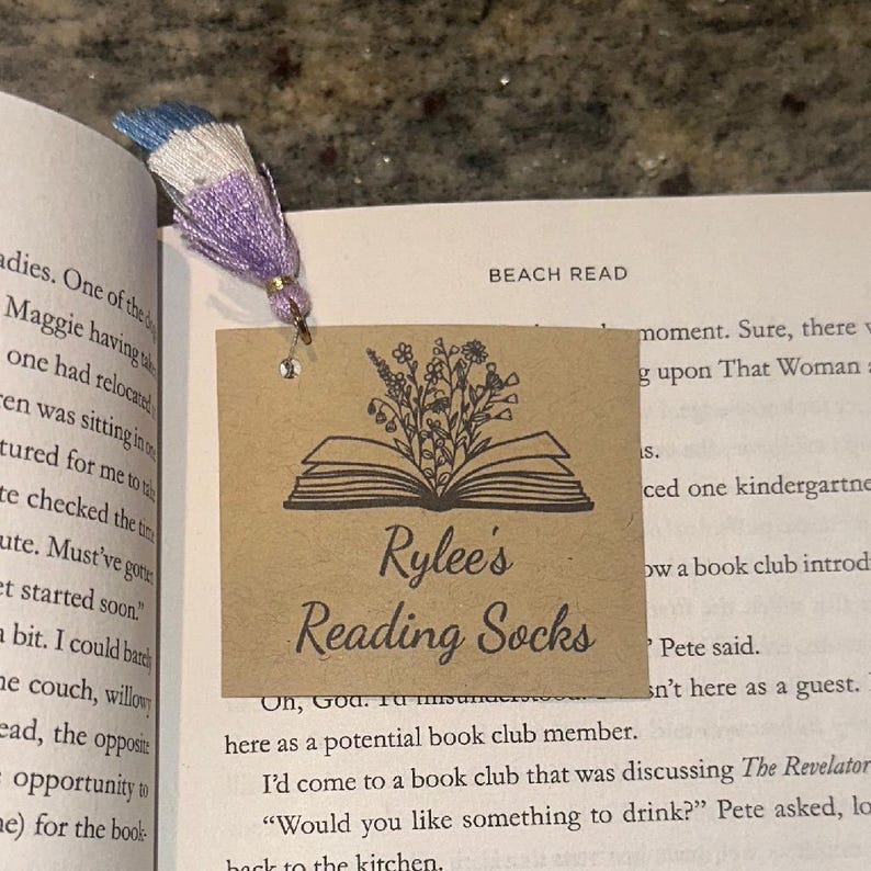 May include: A bookmark with a tassel and a decorative tag. The tag is brown with an illustration of an open book and flowers. The text "Rylee's Reading Socks" is printed on the tag. The tassel is blue, white, and purple.