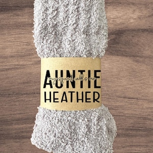 May include: A pair of light gray fuzzy socks wrapped with a brown paper band. The band features the text "AUNTIE HEATHER" in black, with "my squad calls me" above it.