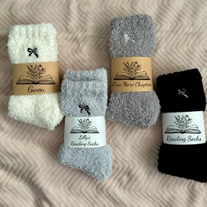 May include: Four pairs of cozy reading socks in white, gray, and black, each with a small bow detail. The socks are wrapped with paper bands featuring an open book and floral design, with text that reads "Gwen", "One More Chapter", and "Lilly's Reading Socks".
