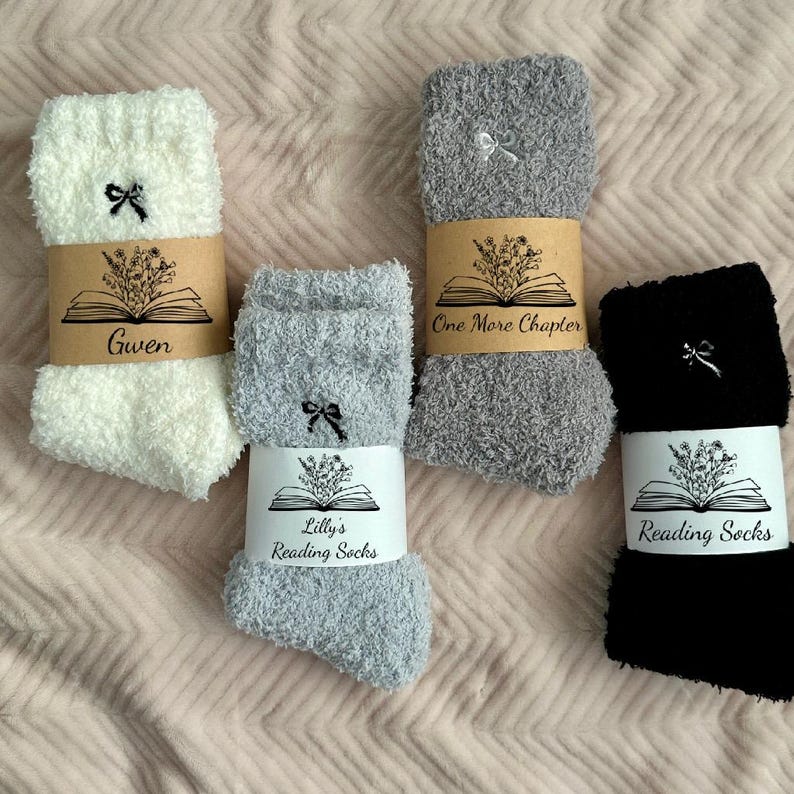 May include: Four pairs of cozy reading socks in white, gray, and black, each with a small bow detail. The socks are wrapped with paper bands featuring an open book and floral design, with text that reads "Gwen", "One More Chapter", and "Lilly's Reading Socks".