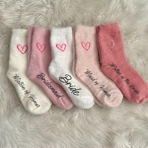 May include: Five pairs of fuzzy white and pink socks with hearts and the words "Bride", "Bridesmaid", "Maid of Honor", "Matron of Honor", and "Mother of the Bride" written on them.