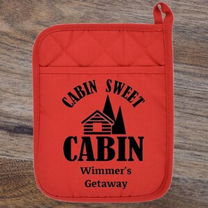 May include: Red quilted oven mitt with black text that reads "Cabin Sweet Cabin Wimmers' Getaway". The text is surrounded by a cabin and pine trees.