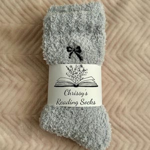May include: A pair of light gray fuzzy socks with a black bow near the top. A white band around the socks features a floral illustration above the text "Chrissy's Reading Socks". The socks are displayed on a pink surface.