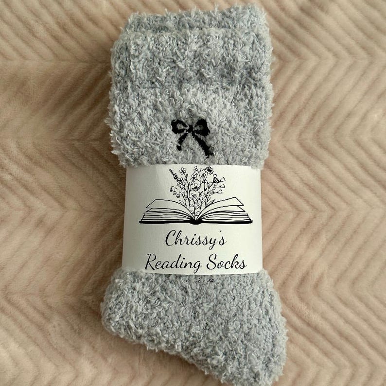May include: A pair of light gray fuzzy socks with a black bow near the top. A white band around the socks features a floral illustration above the text "Chrissy's Reading Socks". The socks are displayed on a pink surface.
