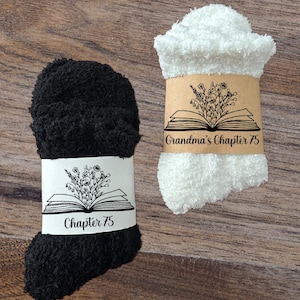 May include: Two pairs of fluffy socks, one black and one white, each wrapped with a band featuring an open book and floral design. The black sock's band reads "Chapter 75," while the white sock's band says "Grandma's Chapter 75."