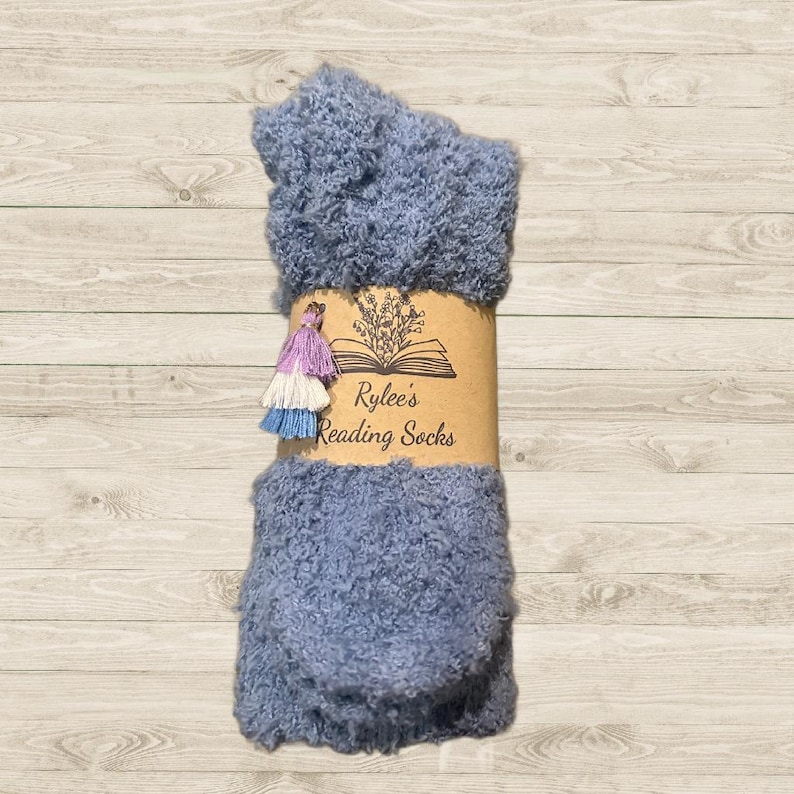 Pu&ograve; includere: Un paio di calzini blu e pelosi con un'etichetta di carta marrone che dice "Rylee's Reading Socks". L'etichetta ha un design floreale e un'illustrazione di un libro aperto.