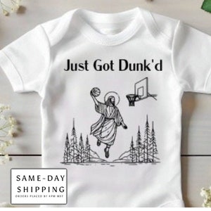May include: White baby onesie with a black and white illustration of a person dunking a basketball. The text "Just Got Dunk'd" is printed in black above the illustration.