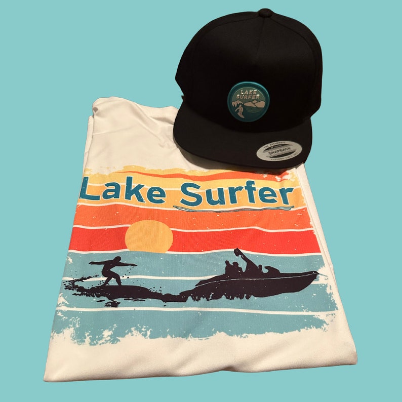 May include: White t-shirt with a retro sunset graphic and the text "Lake Surfer". A black snap-back hat with a turquoise logo that says "Lake Surfer" is also pictured.