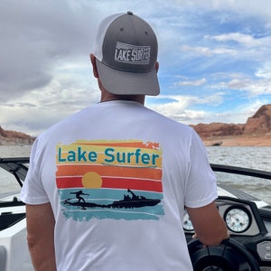 May include: White t-shirt with a retro sunset graphic and the text "Lake Surfer". The graphic features a silhouette of a person waterskiing behind a boat. The person is wearing a life jacket. The t-shirt is being worn by a person wearing a gray mesh trucker hat with the text "Lake Surfer" on the front.