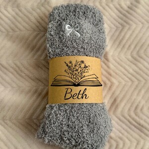 May include: A pair of gray fuzzy socks with a small white bow at the top. A brown paper band wraps around the socks, featuring an open book illustration with flowers and the name "Beth" in script.