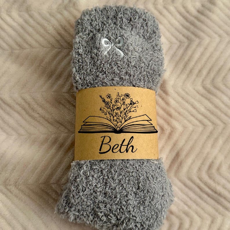 May include: A pair of gray fuzzy socks with a small white bow at the top. A brown paper band wraps around the socks, featuring an open book illustration with flowers and the name "Beth" in script.