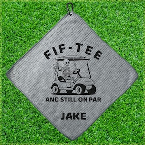 May include: Gray golf towel with a black carabiner clip. The towel features the text "FIF-TEE" above an illustration of a skeleton driving a golf cart, with the text "AND STILL ON PAR" and "JAKE" below.