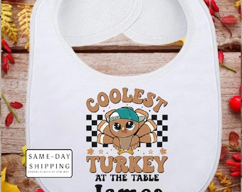 May include: White baby bib with a cartoon turkey wearing a blue cap and the text "COOLEST TURKEY AT THE TABLE James". The bib has a rounded shape and is made of soft material.