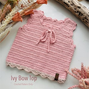 May include: A handmade pink crochet top with a scalloped neckline and hem, featuring a delicate bow detail at the front. The top has a light beige trim and a small brown tag. The text "Ivy Bow Top - Crochet Pattern Only -" is visible.