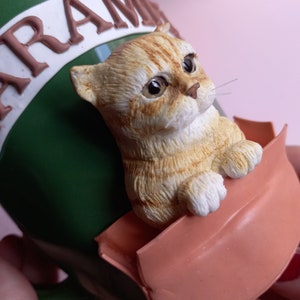 May include: A green ceramic mug with the word "CARAMEL" written on it. A small, orange and white cat figurine is perched on the rim of the mug, holding onto the edge of a pink, flower-shaped handle.