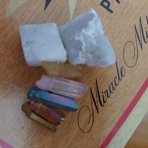 Könnte beinhalten: Eine Sammlung von sechs Kristallen, darunter zwei weiße, ein klarer, ein rosa, ein schillernder und ein goldener, angeordnet auf einer Holzoberfläche mit dem sichtbaren Text "Miracle" und "PAN".