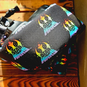 Può includere: Un copriobiettivo per fotocamera nero con una stampa arcobaleno del logo di Star Wars. Il copriobiettivo è in tessuto e ha una chiusura con coulisse.